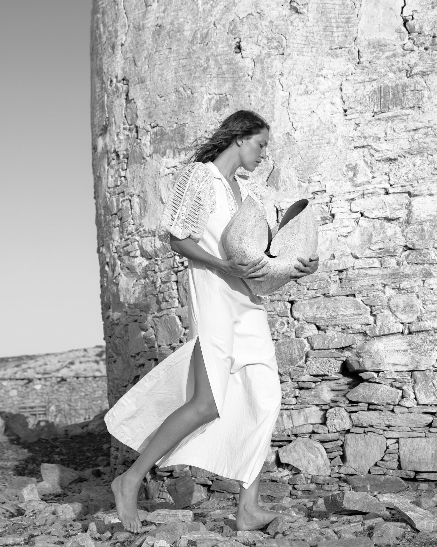 Model walking beside a Paros stone wall holding a pale sculptural ceramic vessel